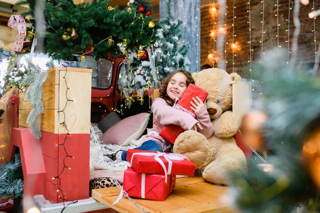 La bambina felice si siede vicino all'abete decorato di Natale e Capodanno e all'orsacchiotto e tiene in mano una grande scatola rossa con un sorriso