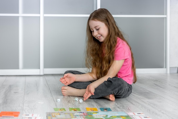 La bambina felice sul pavimento lancia i dadi del gioco per il passatempo del gioco da tavolo