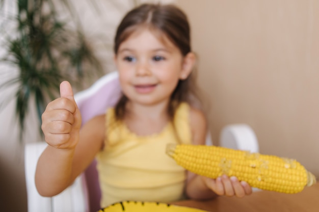 La bambina felice tiene il mais fresco in mano e sorride la ragazzina alza il pollice in alto l'umore estivo a casa focus