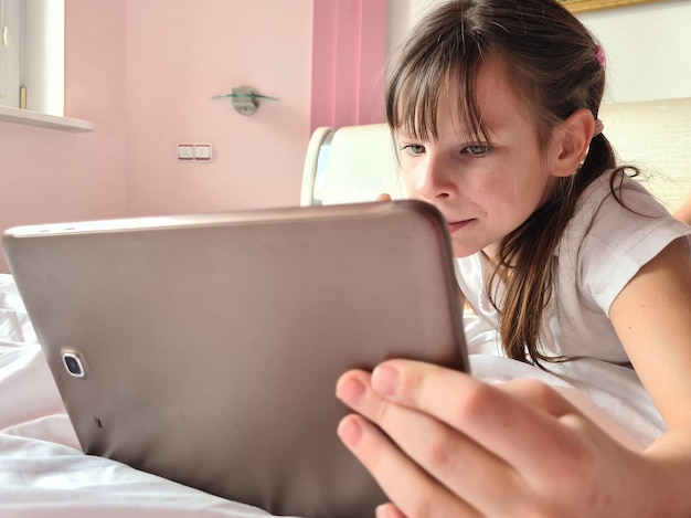 La bambina giace a letto con il tablet e guarda lo schermo del dispositivo