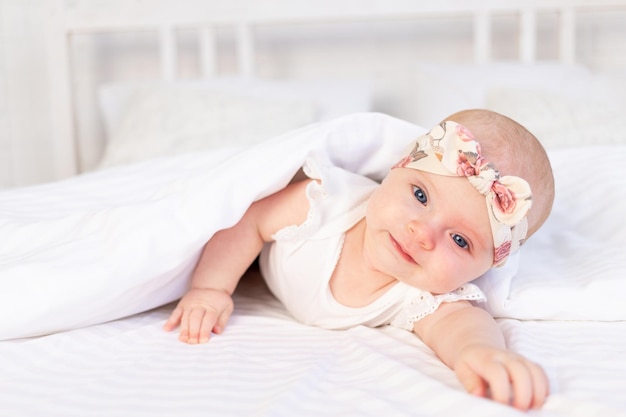 La bambina giace sotto una coperta e sorride su un letto di cotone bianco a casa la mattina del neonato