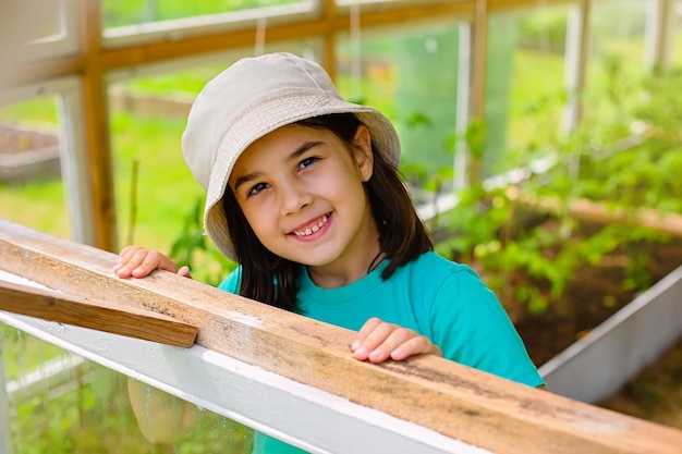 La bambina graziosa in un panama bianco guarda fuori dalla finestra di una serra di verdure in legno