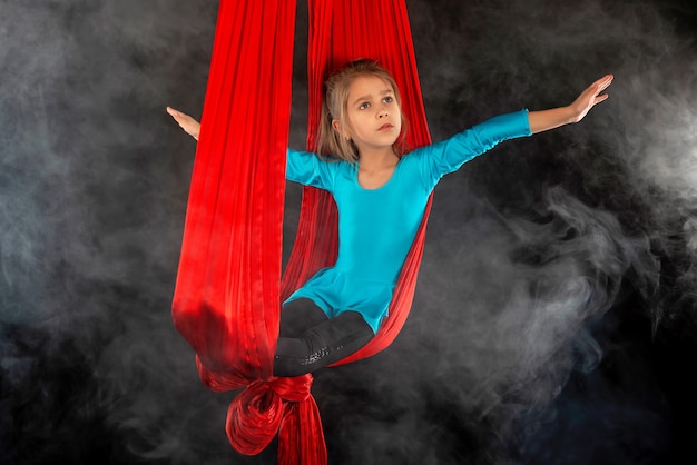 La bambina graziosa senza paura in un vestito da ginnastica blu mostra un nastro rosso aereo acrobatico circondato dal fumo