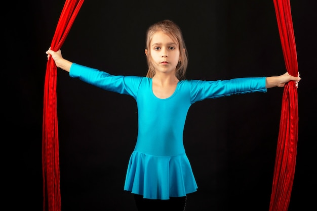 La bambina graziosa senza paura in un vestito da ginnastica blu mostra un nastro rosso aereo acrobatico su un nero