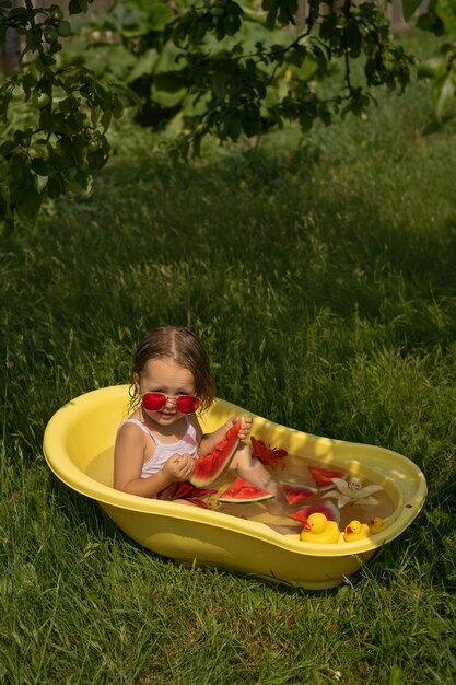 la bambina guarda fuori da dietro gli occhiali da sole rossi mentre mangia un'anguria in una bacinella con fiori