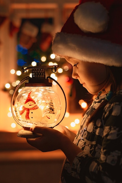 La bambina guarda una lanterna di Natale che illumina il viso con il cappello di Babbo Natale di notte nella stanza