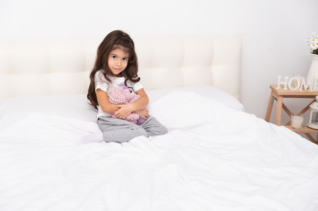 La bambina in abbigliamento domestico si siede con il coniglietto dell'orsacchiotto sul letto a casa.