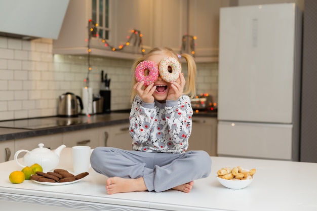 la bambina in abiti di stampa natalizia è seduta in una bella cucina con due ciambelle in mano