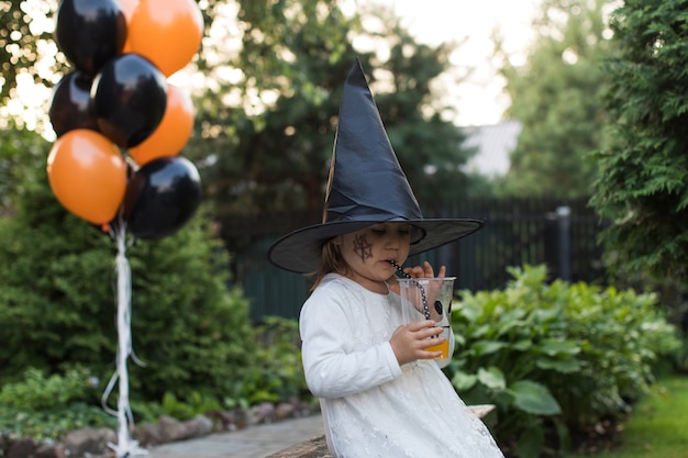 La bambina in abito e cappello da strega beve il succo in cortile