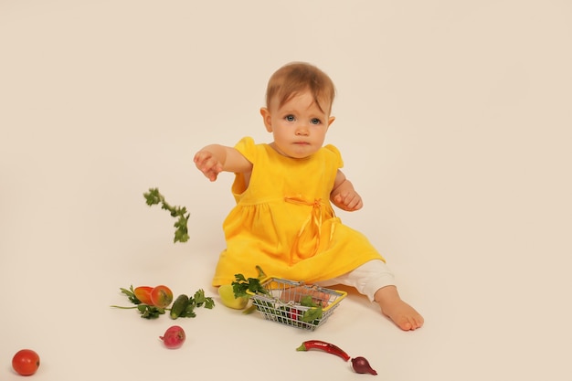 la bambina in abito giallo è seduta su uno sfondo bianco accanto a un piccolo cesto con verdure