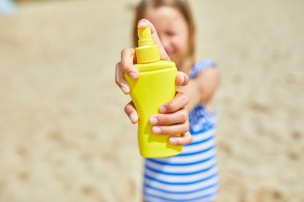 La bambina in costume da bagno tiene in mano una bottiglia gialla di crema solare in piedi sulla spiaggia, protezione della pelle crema solare. Vacanze estive.
