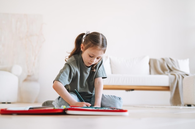 La bambina in età prescolare è seduta sul pavimento e disegna in una stanza luminosa a casa