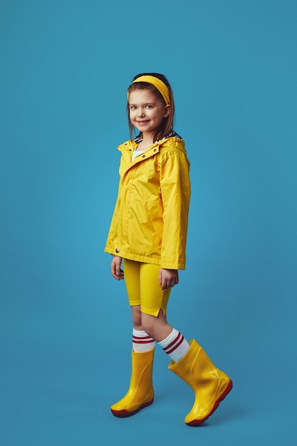 La bambina in impermeabile giallo e scarpe da ginnastica sorride mentre sta in piedi contro