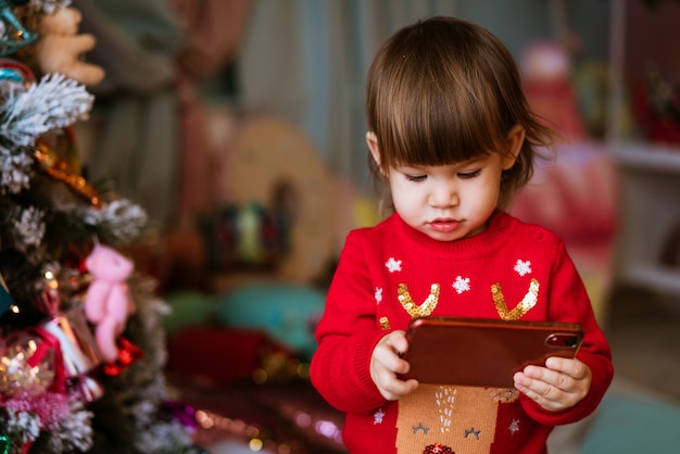 La bambina in maglione rosso sta guardando i cartoni animati sul telefono accanto all'albero di natale a casa mentre si siede...