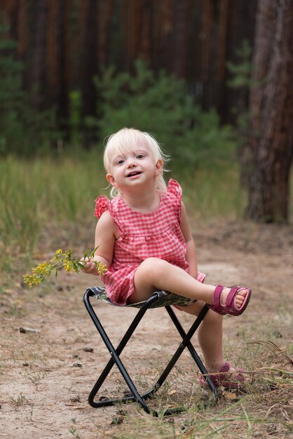 La bambina in prendisole si siede su una sedia contro la foresta