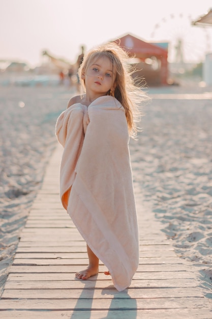 La bambina in un asciugamano sulla spiaggia si trova su un sentiero di legno nella sabbia al tramonto