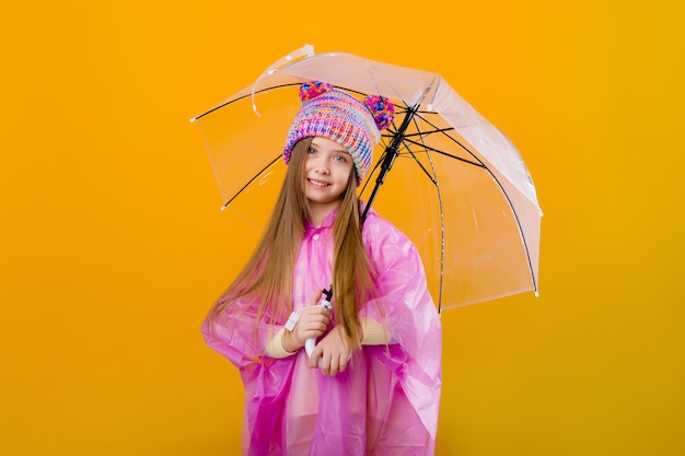 La bambina in un impermeabile rosa e un cappello tricottato sta tenendo un ombrello su uno spazio giallo