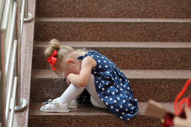 La bambina in un vestito blu si siede sui gradini e piange