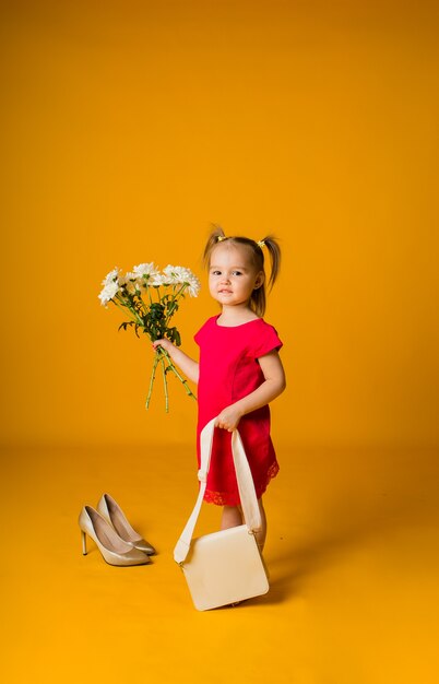 La bambina in un vestito rosso con una borsa beige tiene un mazzo di fiori bianchi su una superficie gialla con spazio per testo