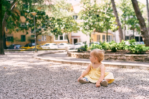 La bambina in un vestito si siede su un sentiero di ghiaia nel cortile della casa