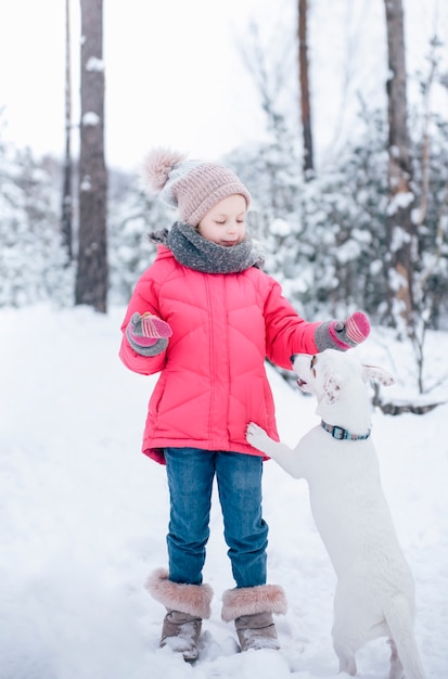 La bambina in una giacca luminosa gioca nella foresta innevata invernale con il suo cane jack russell terrier