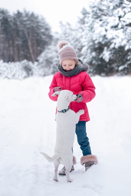 La bambina in una giacca luminosa gioca nella foresta innevata invernale con il suo cane jack russell terrier