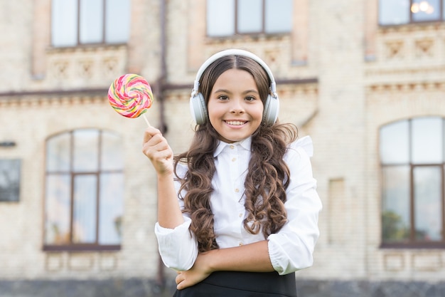 La bambina in uniforme di classe mangia lecca-lecca. di nuovo a scuola. educazione moderna con le nuove tecnologie. bambino al cortile della scuola. immagina che sia cantante. il bambino ha una pausa musicale. ragazza in cuffia.