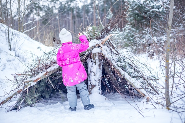 La bambina in vestiti caldi costruisce una capanna dai rami di conifere nella foresta invernale, giocando all'aperto in inverno