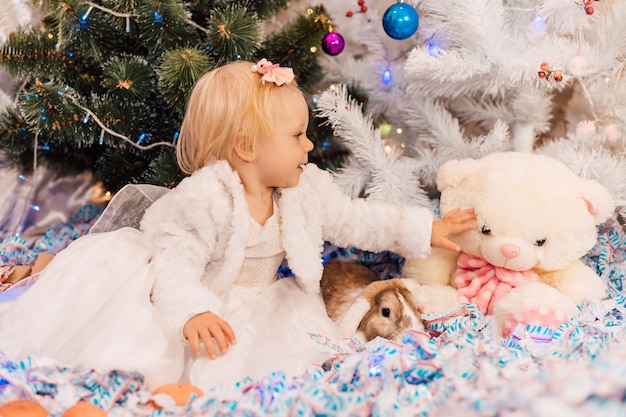 La bambina in vestito bianco gioca vicino all'albero di Natale con un coniglio