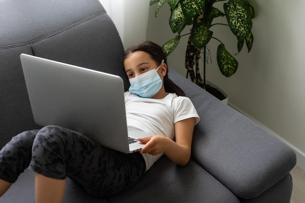 La bambina intelligente che indossa la maschera fa i compiti nel suo soggiorno. È seduta e usa il laptop.