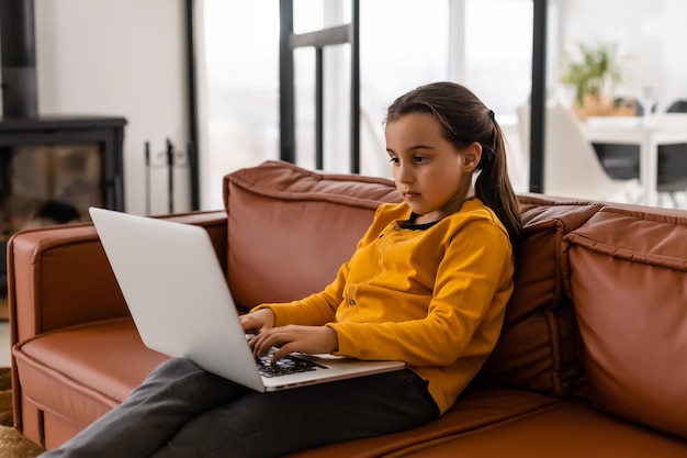 La bambina intelligente fa i compiti nel suo soggiorno. È seduta e usa il laptop.