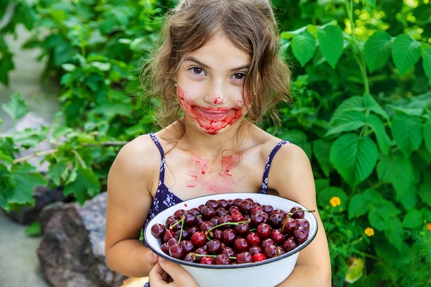 La bambina mangia le ciliegie da una ciotola bianca in giardino con la faccia piena di macchie di ciliegie