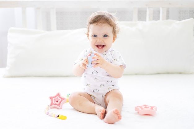 La bambina sana e sveglia in un body bianco è seduta sul letto su lenzuola bianche sorridendo