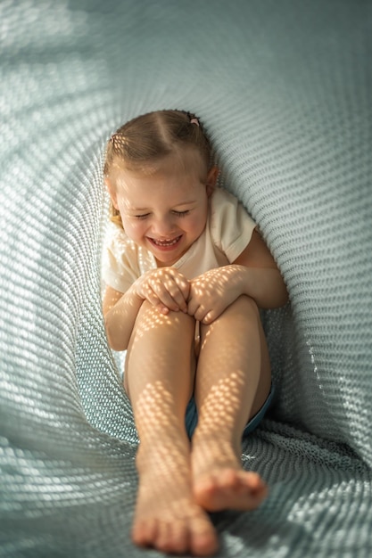 La bambina si diverte sotto il plaid lavorato a maglia blu nella mattinata di sole