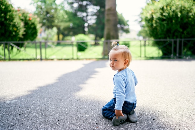 La bambina si siede in ginocchio sull'asfalto sullo sfondo di una recinzione in un parco verde