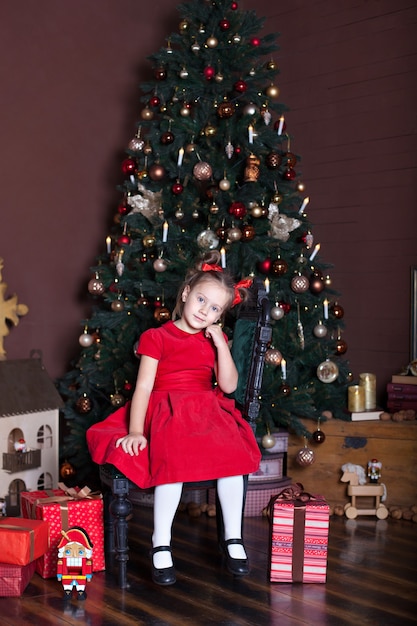 La bambina si siede nella stanza vivente vicino all'albero di Natale e ai regali