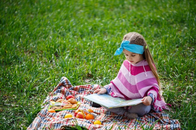 La bambina si siede su un copriletto e legge un libro con una fiaba, erba verde nel campo, tempo primaverile soleggiato, sorriso e gioia del bambino, cielo blu con nuvole