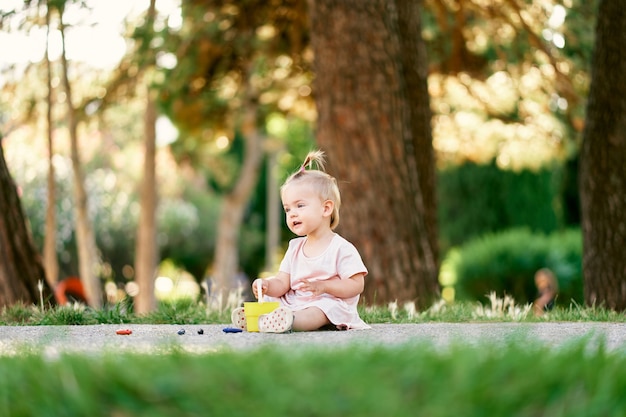 La bambina si siede su un sentiero in un parco verde con un secchio giocattolo in mano