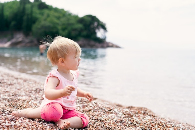 La bambina si siede su una spiaggia di ciottoli vicino all'acqua sullo sfondo di un'isola verde