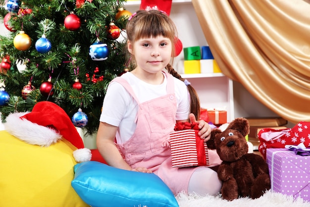 La bambina si siede vicino a un albero di Natale con un regalo in mano in una stanza decorata a festa