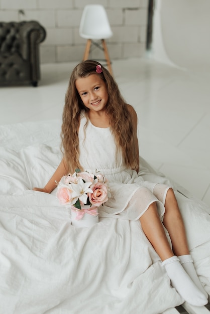 La bambina sorridente con i capelli lunghi si siede su un letto bianco