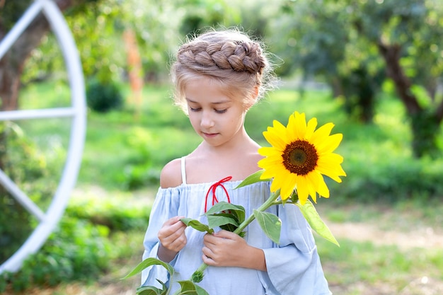 La bambina sorridente con una treccia sulla sua testa tiene il girasole in giardino. Ragazza bionda del ritratto del primo piano giovane con il girasole.