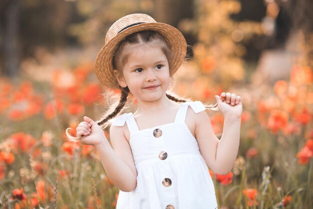 La bambina sorridente di 3-4 anni indossa il cappello di paglia e il vestito rustico bianco che posa sopra il fondo del papavero della natura. Bambino felice nel prato fiorito. Tempo di primavera. Infanzia.