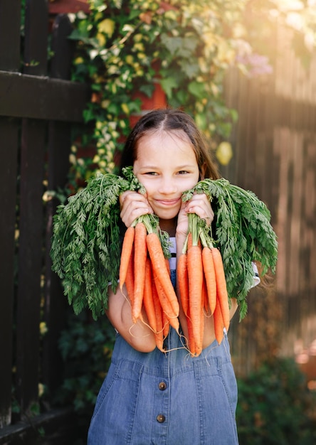 La bambina sorridente in un giardino tiene due mazzi di carote fresche verticali