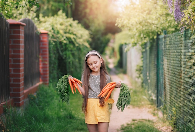 La bambina sorridente in un giardino tiene due mazzi di carote fresche