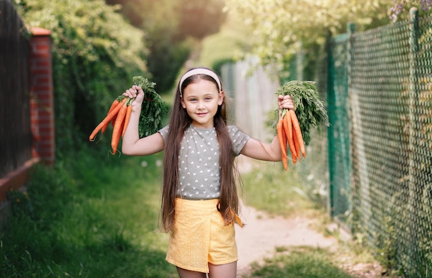 La bambina sorridente in un giardino tiene due mazzi di carote fresche