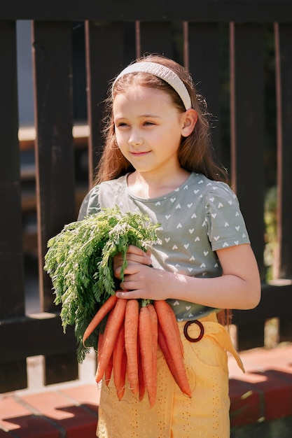 La bambina sorridente in un giardino tiene un mazzo di carote fresche Verticale