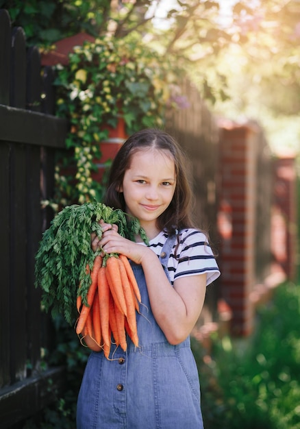 La bambina sorridente in un giardino tiene un mazzo di carote fresche Verticale