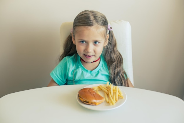 La bambina sorridente si siede al tavolo con un piatto di hamburger e patatine fritte