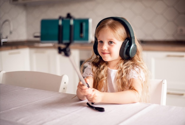 La bambina sorridente si siede sul divano in soggiorno indossa le cuffie guarda il video sul cellulare il bambino felice in cuffia si rilassa sul divano si diverte utilizzando lo smartphone a casa i bambini e il concetto di tecnologia